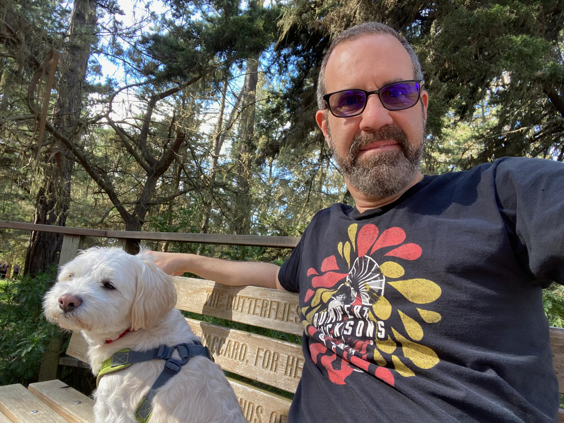 Tom Cohen, in sunglasses and a gray T-shirt with red and yellow petals on it, sitting on a bench with a small white dog