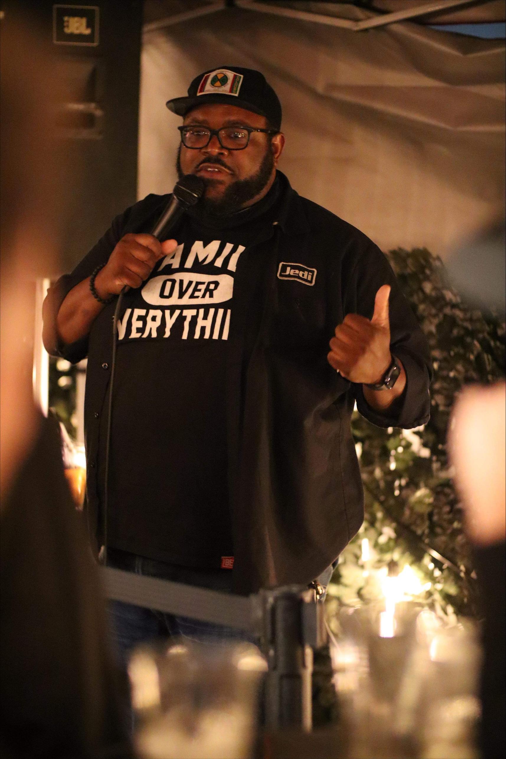 Ato Walker, in a black hat and shirt, standing in a dimly lit room with a microphone